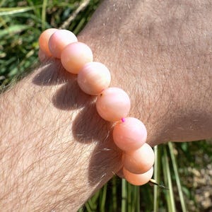 May include: A bracelet made of peach-colored, round beads. The bracelet is worn on a person's wrist, with the beads appearing to be approximately 0.75 inches in diameter. The background is blurred, showing green grass.