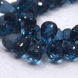 May include: Close-up of a strand of faceted, teardrop-shaped gemstones in varying shades of deep blue. The gems are arranged in a circular pattern, with light reflecting off their cut surfaces. The background is a plain white surface.