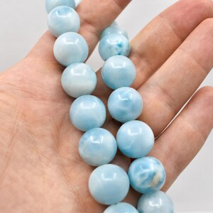 May include: A strand of light blue, round gemstone beads. The beads are held in a hand, showcasing their smooth, polished surface and varying shades of blue. The beads are approximately 10mm in diameter.