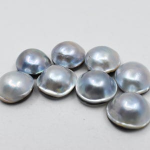 Peut inclure: Huit perles bleu-gris iridescentes en forme de dôme sont disposées sur une surface blanche. Les perles ont une surface lisse et réfléchissante, avec une subtile variation de couleur. Ces perles sont idéales pour la création de bijoux.