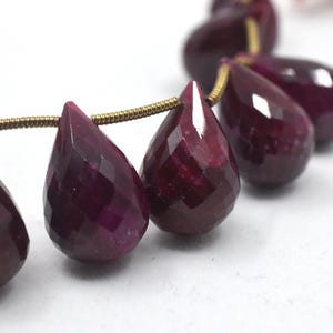 May include: A strand of faceted, teardrop-shaped gemstones in a deep burgundy color. The gems are strung on a gold-colored wire, creating a necklace or jewelry component. The stones have a polished, reflective surface.