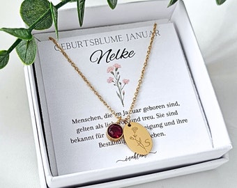 May include: A gold necklace with a red crystal charm and a round pendant engraved with a flower and the letter "S". The necklace is in a white box with a card that reads "Geburtsblume Januar Nelke" and a description of the flower's meaning.