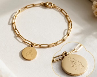 May include: A gold-toned chain link bracelet featuring a circular pendant. The pendant is engraved with a line art illustration of a hand making a finger heart gesture with a small heart symbol above it. The bracelet has a lobster claw clasp.