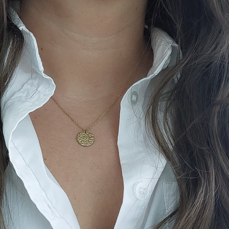 May include: A gold necklace with a hammered round pendant. The necklace is worn by a person with long brown hair and a white collared shirt.