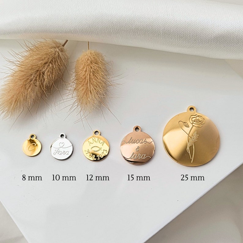 K&ouml;nnte beinhalten: F&uuml;nf goldfarbene Metallscheiben mit verschiedenen Gravuren. Die Scheiben haben einen Durchmesser von 8 mm, 10 mm, 12 mm, 15 mm und 25 mm. Die Gravuren beinhalten ein Herz, einen Fisch, die Namen "Lucas & Lora" und eine Blume.