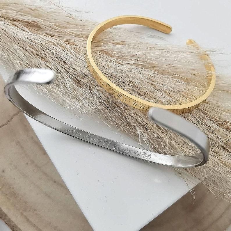 Op de afbeelding: Twee gouden en zilveren armbandjes met gegraveerde tekst. De gouden armband zegt "Happiness" en de zilveren armband zegt "My heart is yours".