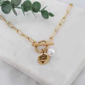 Op de afbeelding: Gouden ketting met een toggle-sluiting en een bedel met een hart en de naam "Louis" gegraveerd op een ronde schijf. Een witte parel hangt aan de bedel.