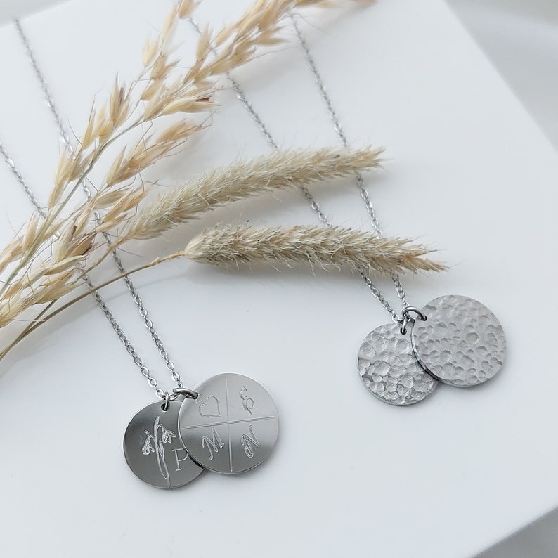May include: Two silver necklaces with round pendants. One pendant has a flower design and the letters "F M" and "&" engraved on it. The other pendant has a textured surface.