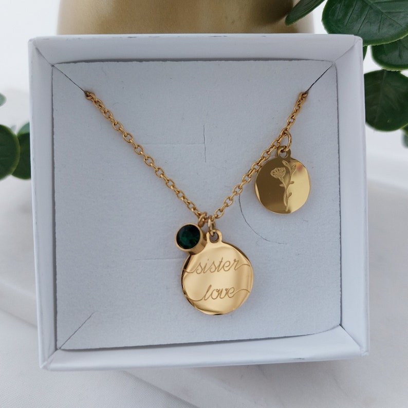 May include: Gold necklace with two round pendants. One pendant has the words "sister love" engraved on it and the other has a small flower design. A green gemstone is attached to the chain above the "sister love" pendant.