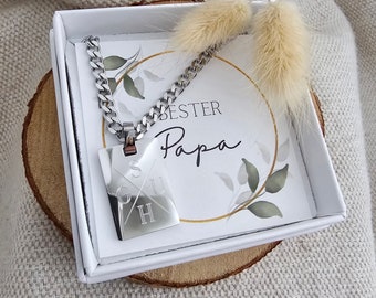 Op de afbeelding: Een zilveren ketting met een vierkante hanger waarop "SUCH" in een kruis staat. De ketting zit in een witte doos met een kaart waarop "BESTER PAPA" staat met een bloemenontwerp.