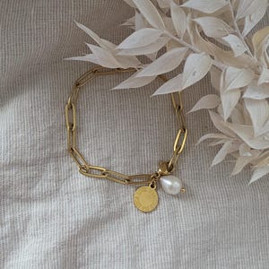 May include: Gold-colored chain bracelet with a pear-shaped pearl charm and a small round charm. The bracelet is made of linked oval segments. The round charm has text on it. The bracelet is displayed on a neutral-colored fabric.