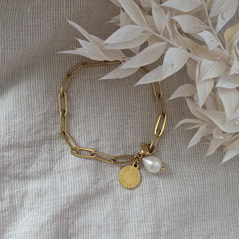 May include: Gold-colored chain bracelet with a pear-shaped pearl charm and a small round charm. The bracelet is made of linked oval segments. The round charm has text on it. The bracelet is displayed on a neutral-colored fabric.