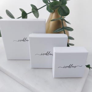 May include: Three white gift boxes of varying sizes are arranged on a marble surface. Each box has the word "iraklam" printed in a delicate script. A gold vase and green foliage are in the background.