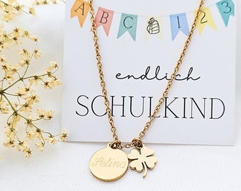 Op de afbeelding: Gouden ketting met een ronde bedel gegraveerd met de naam "Selina" en een klavertje vier bedel. De ketting ligt op een witte kaart met de tekst "endlich SCHULKIND" en kleurrijke vlaggen met letters en cijfers.