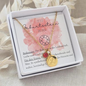 Könnte beinhalten: Goldfarbene Halskette mit einem kleinen roten Edelstein und einem runden Anhänger mit dem eingravierten Buchstaben "L". Die Halskette wird in einer weißen Schachtel mit einer Karte mit deutschem Text und Aquarellhintergrund präsentiert.