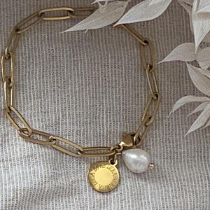 May include: Gold-toned chain bracelet with a large link design. The bracelet features a round charm with engraved text and a white pearl drop. The bracelet is displayed on a neutral linen surface.