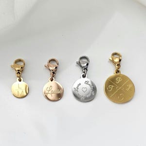 K&ouml;nnte beinhalten: Vier goldene, silberne und ros&eacute;goldene Anh&auml;nger mit verschiedenen Gravuren. Die Anh&auml;nger zeigen ein Herz, Initialen und einen Namen. Die Anh&auml;nger sind an einem Karabinerhaken befestigt.