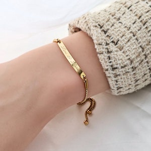 Pode incluir: Pulseira ajustável de ouro com um charme retangular fino gravado com o texto "Love & Live".
