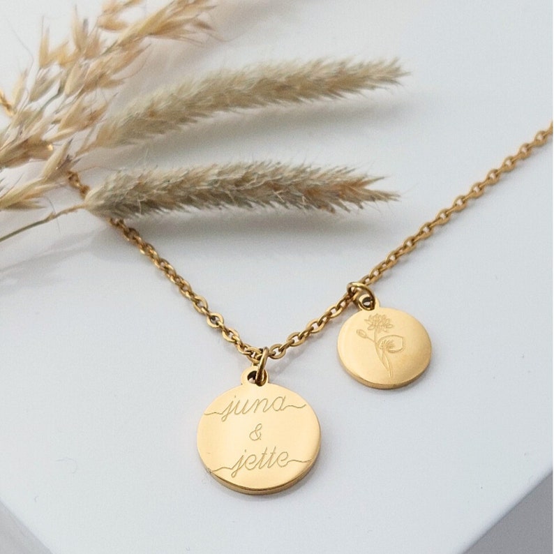 May include: Gold necklace with two round pendants. One pendant has the text "juna & jette" engraved on it. The other pendant has a delicate floral design.