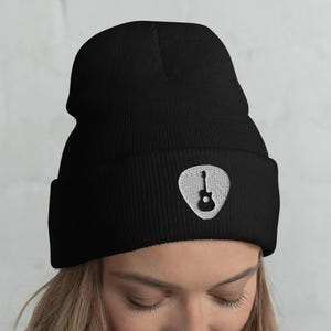 May include: Black beanie hat with a white embroidered guitar pick design. The hat is made of a soft, knitted material and has a folded brim. The guitar pick design features a guitar silhouette. The hat is a stylish accessory for musicians or music lovers.