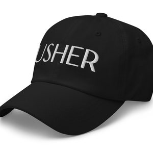May include: Black baseball cap with the word "USHER" embroidered in white across the front. The cap has a curved brim and a fabric strap closure on the back.