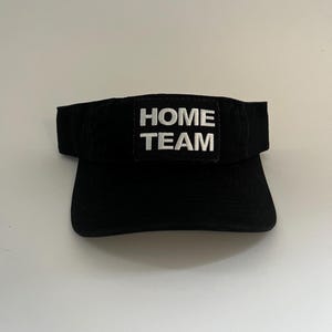 May include: Black visor with the text "HOME TEAM" embroidered in white. The visor has a curved brim and a solid black band. The text is centered on a rectangular patch.