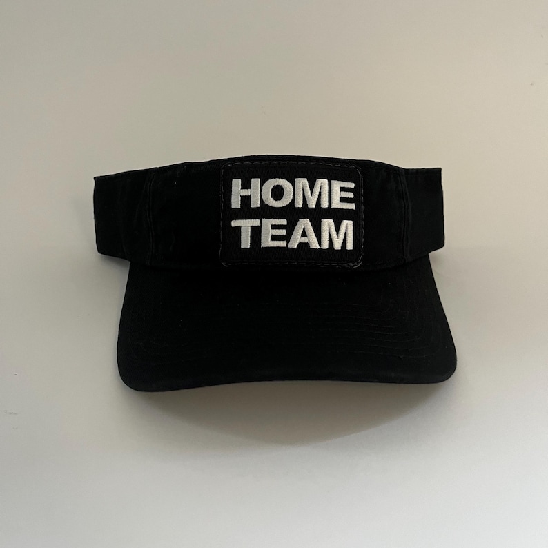 May include: Black visor with the text "HOME TEAM" embroidered in white. The visor has a curved brim and a solid black band. The text is centered on a rectangular patch.