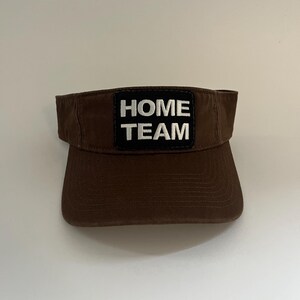 May include: A brown visor with a curved peak and a black patch. The patch displays the words "HOME TEAM" in white, block letters. The visor is made of a durable fabric and is designed to shield the eyes from the sun.
