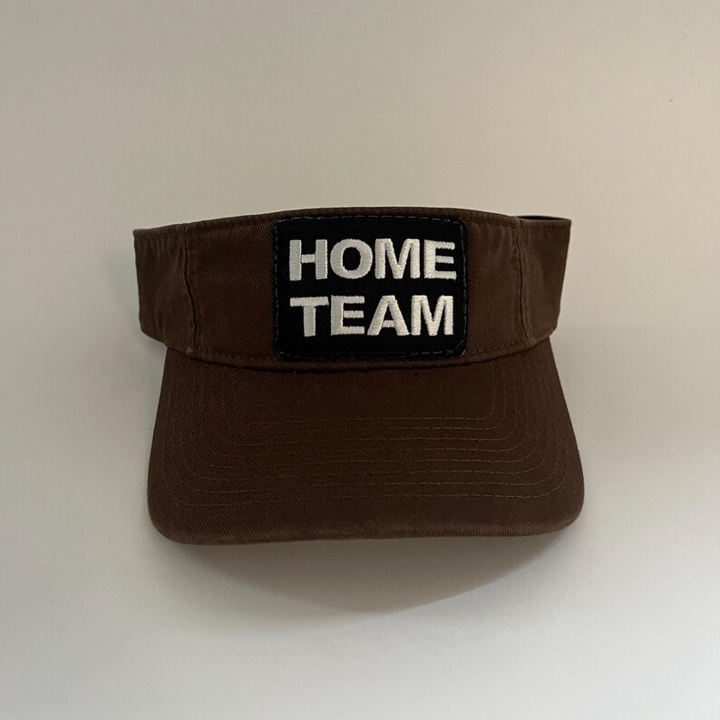 May include: A brown visor with a curved peak and a black patch. The patch displays the words "HOME TEAM" in white, block letters. The visor is made of a durable fabric and is designed to shield the eyes from the sun.