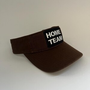 May include: A brown visor with a curved peak. A rectangular patch on the front displays the words "HOME TEAM" in white block letters. The visor is made of a textured fabric.