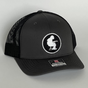 May include: A charcoal gray trucker hat with a black mesh back. The hat features a white embroidered patch with a musician playing an instrument. The Richardson 112 Original Trucker hat is adjustable.