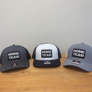 May include: Three baseball caps featuring a rectangular patch on the front with the text HOME TEAM in white letters on a black background. The caps are charcoal gray, white with a black brim, and light gray, each with a mesh back and a small label.