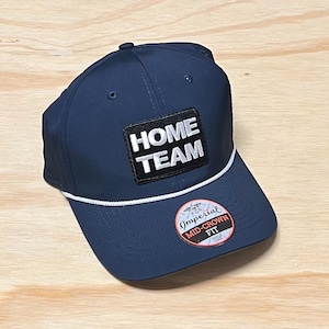 May include: A navy blue baseball cap with a white rope trim and a black patch that says "HOME TEAM" in white lettering. The cap also has a white label that says "Imperial Mid-Crown Fit" in black lettering.