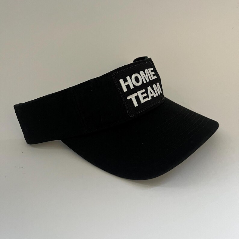 May include: Black visor featuring the text "HOME TEAM" in white. The visor has a curved peak and a soft, matte texture. The text is on a rectangular patch. The visor is designed to shield the eyes from the sun.