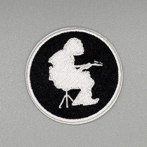 May include: Round black and white patch featuring a musician playing a guitar while seated on a stool. The musician and stool are white, set against a black background. The patch has a white border.
