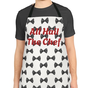 Pode incluir: Um avental branco com um padrão de gravata borboleta preta e o texto vermelho "All Hail The Chef". O avental é usado por uma pessoa e tem uma alça de pescoço preta e laços na cintura.