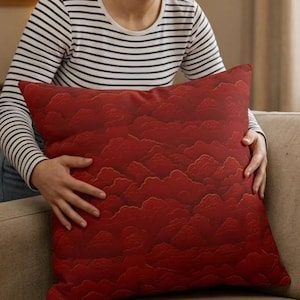 May include: A square, red decorative pillow with a cloud-like pattern. The pillow is held by a person wearing a black and white striped long-sleeve shirt. The pillow is on a beige couch.