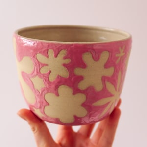 May include: A pink ceramic bowl with a beige floral pattern. The bowl is held by a hand against a light pink background. The interior of the bowl is beige.