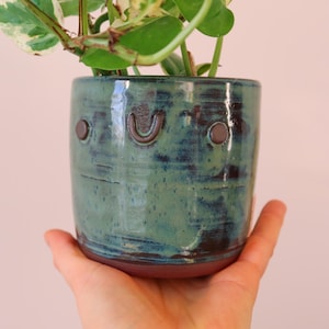 May include: A teal and brown ceramic plant pot, held in a hand. The handmade cylindrical pot has a textured surface and a face design. The pot is filled with a green and white leafy plant. The pot is approximately 10 cm in height.