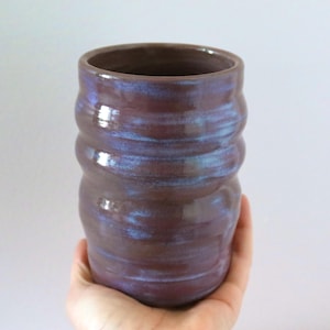 May include: A cylindrical ceramic tumbler with a deep purple glaze and subtle blue highlights. The vessel features a series of horizontal ridges along its body, creating a textured, undulating surface with a glossy finish and a wide, open rim.