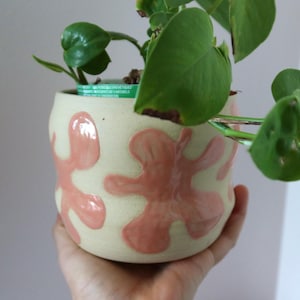 May include: A cylindrical ceramic planter with a matte cream-coloured exterior featuring raised, glossy, pink-coloured organic blob shapes. A green plastic nursery pot is visible inside the planter, containing a green leafy plant with several stems.