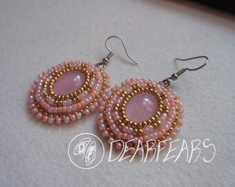 May include: A pair of handmade oval earrings. Each earring features a central pink stone surrounded by layers of pink and gold beads. The earrings have silver-colored ear wires. The text "DEARFEARS" is visible.