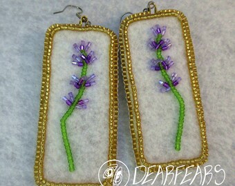 May include: Handmade rectangular earrings featuring a beaded lavender flower design. The earrings have a gold beaded border and a white background. The lavender flowers are purple and green, with a green stem. The earrings are a unique accessory.