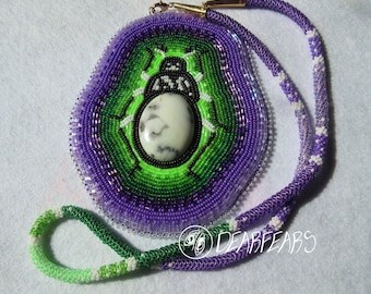 May include: A beaded necklace with a green and purple pendant featuring a black and white beetle design with a white stone in the center. The pendant is surrounded by a purple and green beaded border. The necklace is made of purple and green beads with white accents.