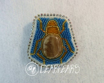 May include: A beaded scarab beetle brooch with a brown stone center, surrounded by blue and gold beads. The brooch is framed by a border of clear iridescent beads.