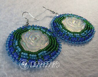 May include: A pair of handmade beaded earrings. Each earring features a central iridescent white rose surrounded by concentric circles of blue and green beads. The earrings have silver-colored ear wires and the text "DEARFEARS" is visible.