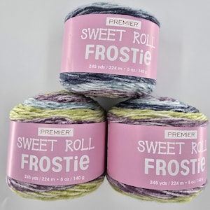 May include: Three skeins of Premier Sweet Roll Frostie yarn. Each yarn ball is wrapped with a pink label that reads "SWEET ROLL FROSTIE" in white text. The yarn is a mix of colors, including blue, green, purple, and white. Each skein contains 245 yards and 5 oz.