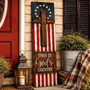 May include: A tall rectangular wooden sign featuring a painted American flag design with a circle of thirteen white stars on a blue field. A dark brown wooden cross is mounted on the center. The text reads THIS IS God's COUNTRY in white.