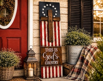 May include: A tall rectangular wooden sign featuring a painted American flag design with a circle of thirteen white stars on a blue field. A dark brown wooden cross is mounted on the center. The text reads THIS IS God's COUNTRY in white.