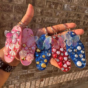 May include: A collection of keychains in pink, blue, and red, each adorned with butterfly accents and sparkling embellishments. The keychains feature various shapes and are decorated with rhinestones, pearls, and floral details.